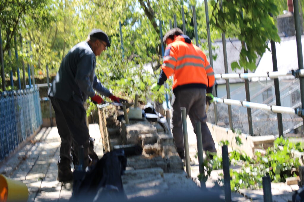 Two workers in a safety-vested orange jacket and dark clothing lean over a railing on a sunny, tree-lined walkway.