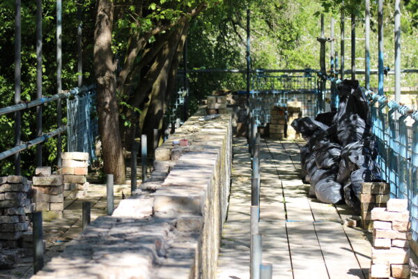 Narrow walkway along a construction site with brick walls, metal railings, and stacked debris bags on the right.