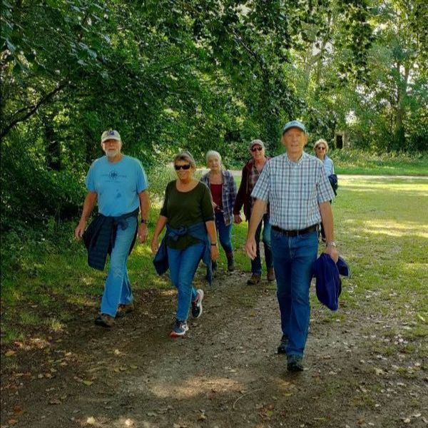 Group of six seniors walking along a dirt path in a shaded park, jackets tied around waists.