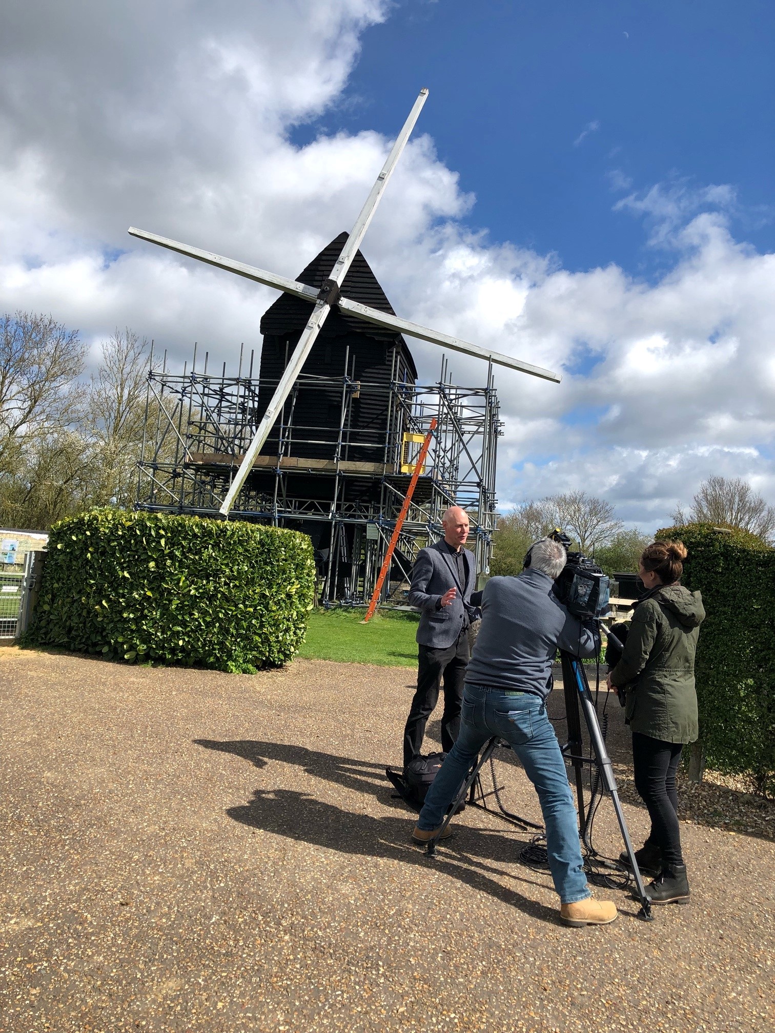 News crew filming outside a small black wooden church under scaffolding, with a large white cross-shaped beam resting across the roof.