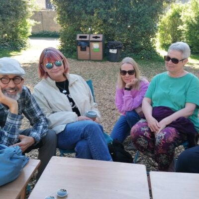 Four adults sitting outdoors around a small table, chatting, with trees and recycling bins in the background.