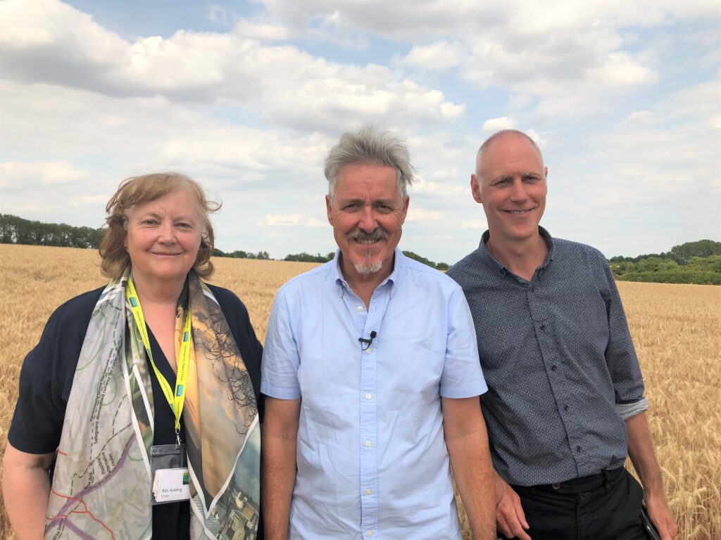 Three adults posing outdoors in a golden field, smiling at the camera under a blue sky with clouds.