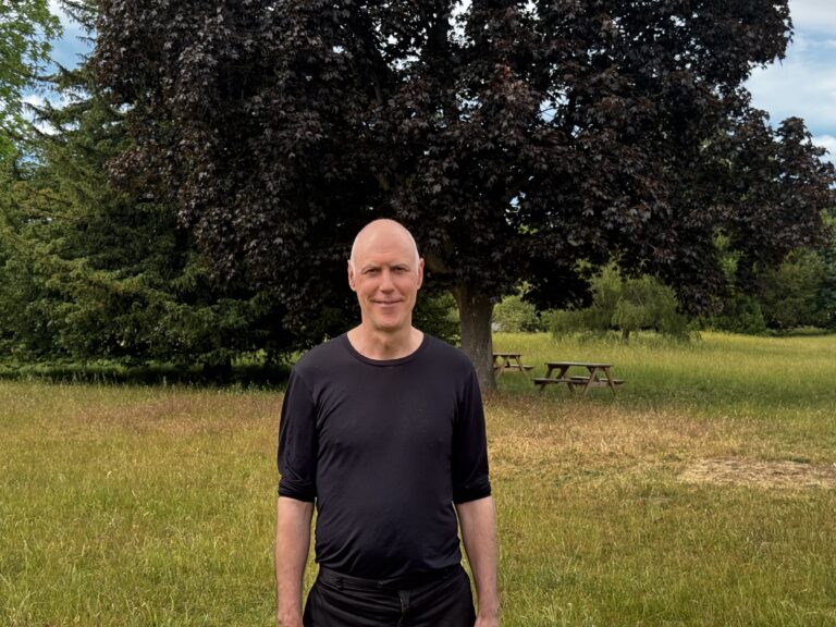 Bald man in a black long-sleeve shirt stands in a grassy park with a large dark-leaved tree behind him and picnic tables in the background.