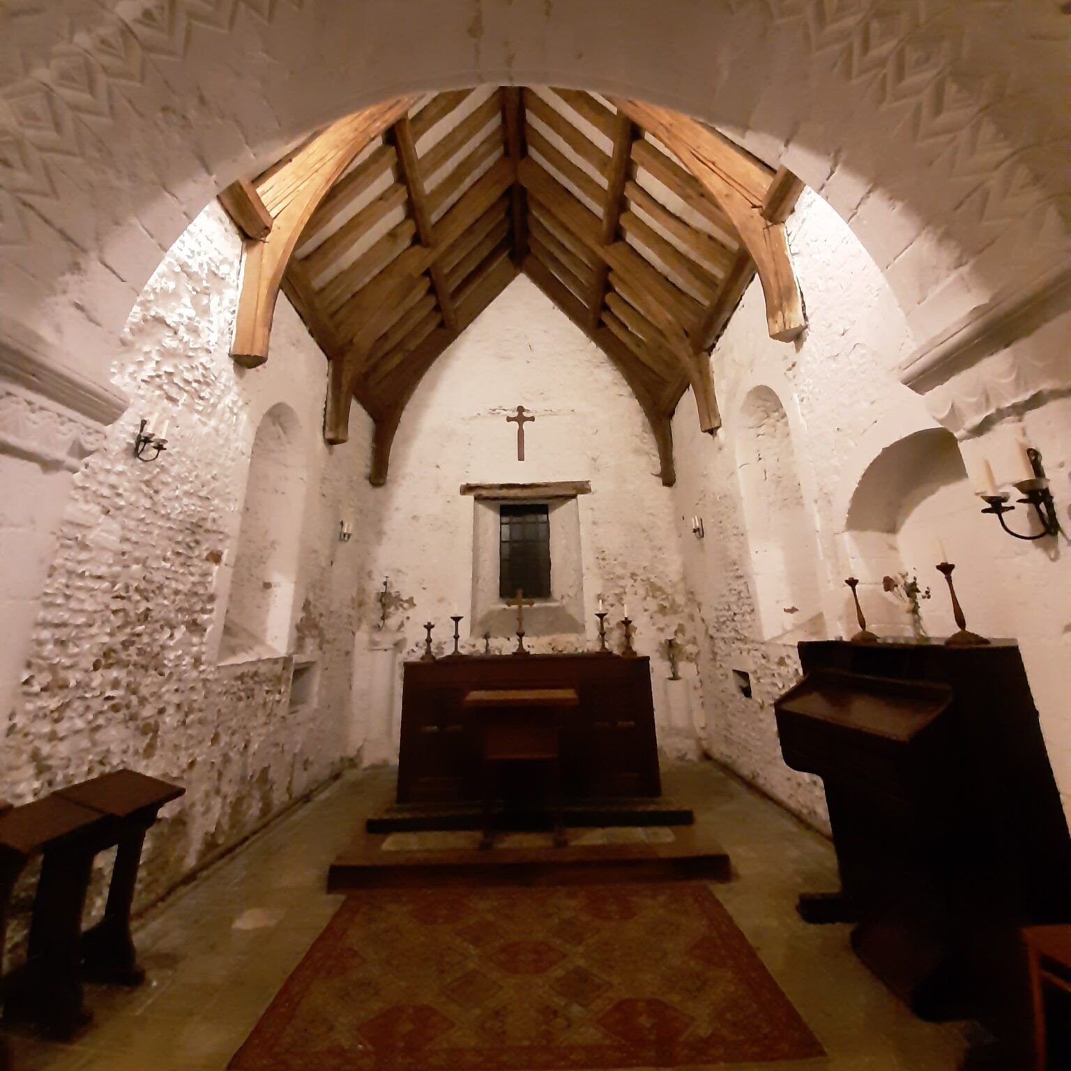 Interior of a small stone church with a wooden vaulted ceiling, a cross above the altar, and candles along the walls.