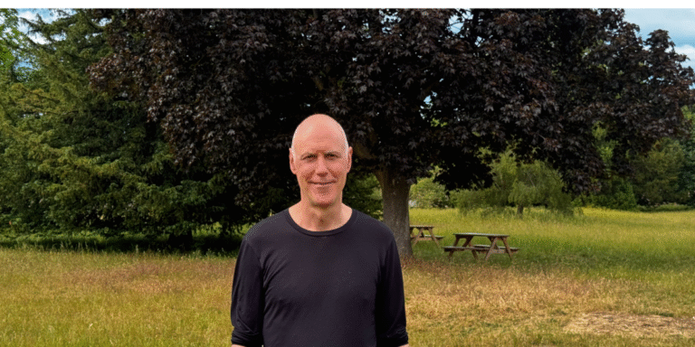Smiling man in a black shirt standing in a grassy park with a large tree and picnic tables in the background.