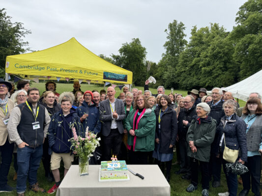 Large group of people smiling and posing outdoors in front of a bright yellow tent that reads 'Cambridge Past, Present & Future'. On a table, a decorated cake with candles and a vase of flowers are visible as attendees celebrate.