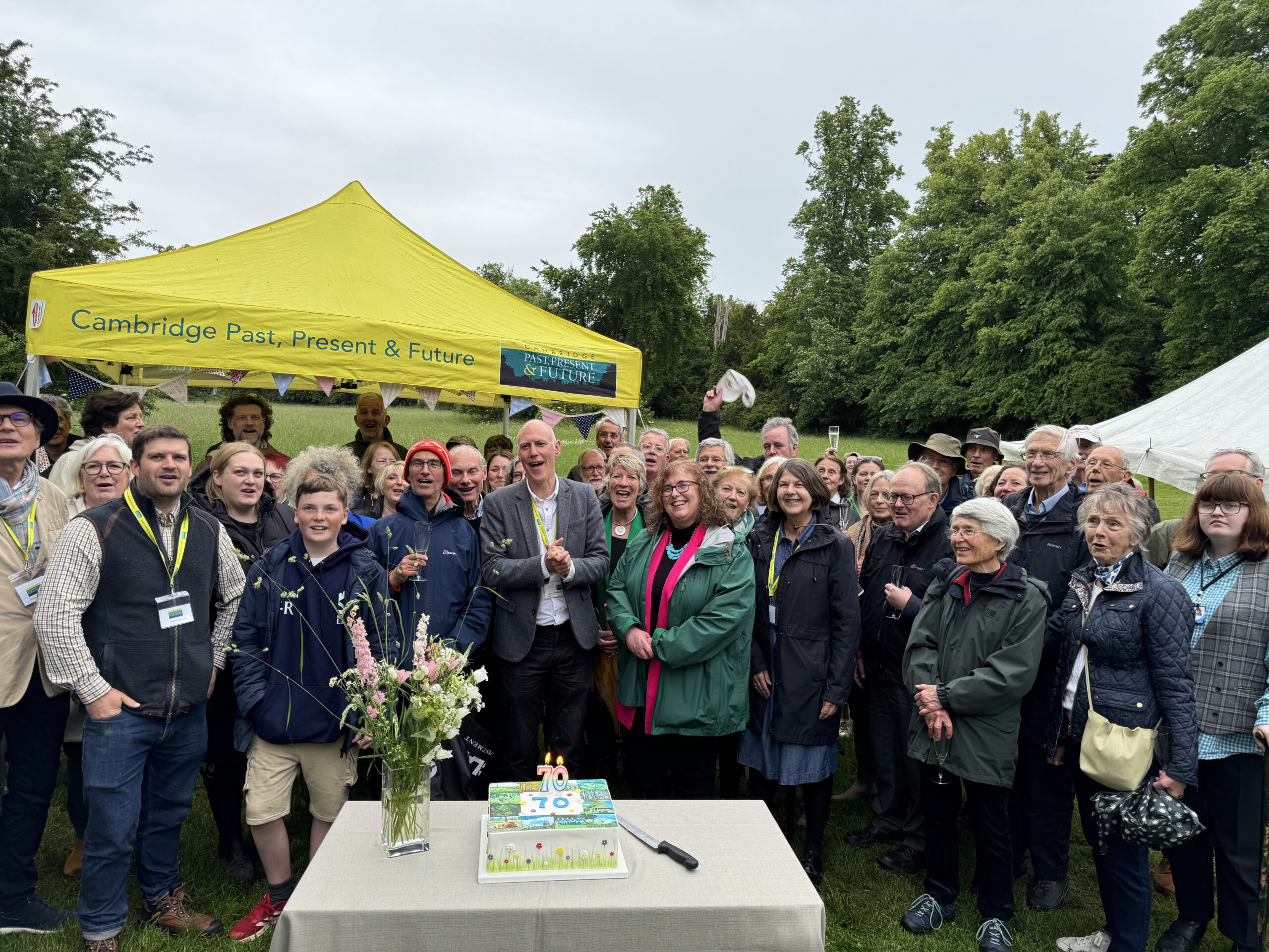 Large group of people smiling and posing outdoors in front of a bright yellow tent that reads 'Cambridge Past, Present & Future'. On a table, a decorated cake with candles and a vase of flowers are visible as attendees celebrate.