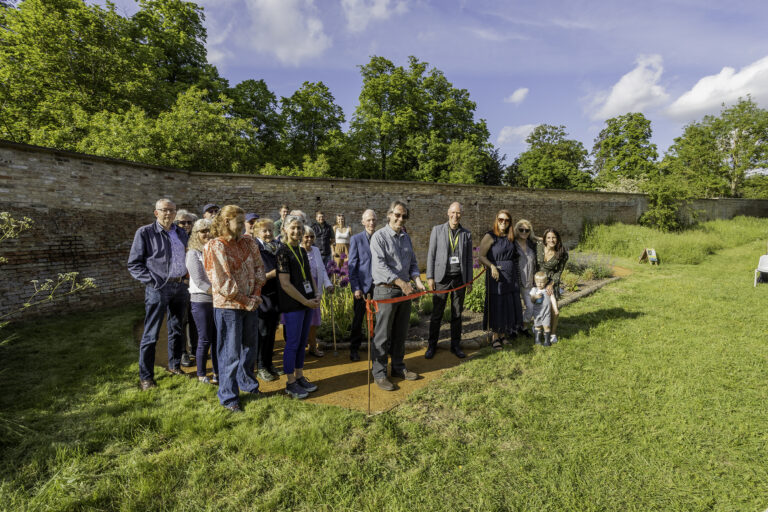 Group of people posing outdoors for a ribbon-cutting ceremony near a brick wall with trees in the background.