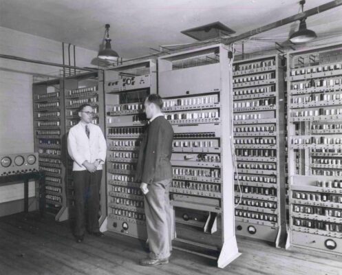 Two technicians stand near rows of rack-mounted computer modules in a vintage data room with large wiring and control panels behind them.