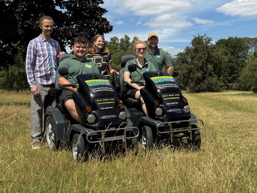 Five adults posing outdoors beside two black mobility scooters with banners on a sunny day in a grassy field.