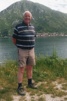 Older man smiling and posing by a lake with green, mountainous backdrop behind him.