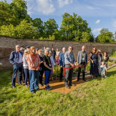 Group of adults and children at a ribbon-cutting ceremony in a garden beside a brick wall.