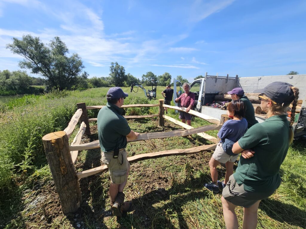 Group of people outdoors by a wooden fence, near a truck with stacked logs, listening to a man in a maroon shirt under a blue sky.