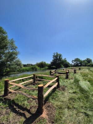 Wooden split-rail fence runs along a grassy path beside a narrow pond under a clear blue sky.