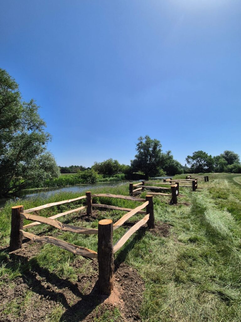 Wooden split-rail fence runs along a grassy path beside a narrow pond under a clear blue sky.
